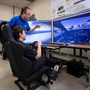 Professor Guoyan Wu teaching a student through a car simulation test. The student is sitting in a chair in front of 2 screens that are displaying the car simulation on-screen.
