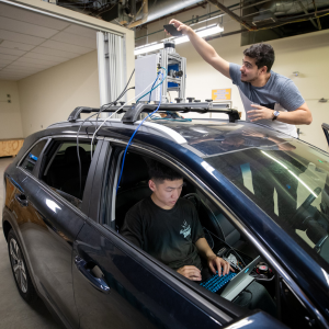 2 members of the UCR EcoCar Team working at CE-CERT. One person is inside the car and a second person is above the car working on a piece of equipment.